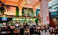 The bar area inside The Treasury, showing large stone pillars, large plants, a large wall clock and lots of bottles of drink displayed.