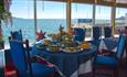 A table inside the restaurant set for a meal with dishes of food in front of large glass windows overlooking the water of Plymouth Sound.