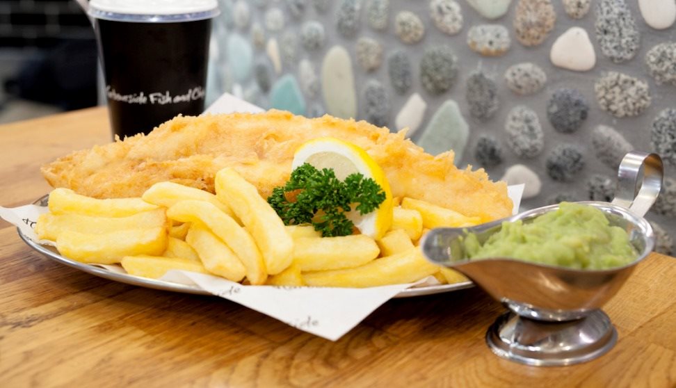 A plate of fish and chips garnished with lemon and parsley and a jug of mushy peas.
