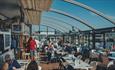 Inside the Boathouse Café with the roof retracted. Customers sitting at tables with a blue sky and views over Sutton Harbour.