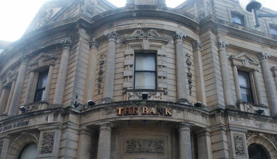 The stone exterior of The Bank with stone pillars and arches and a metal sign saying The Bank.