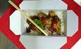 White cardboard takeaway box containing a meal and rice and a pair of chopsticks balanced inside.