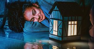 A woman lies with her head on her arm. She smiles, looking at a model of a house which is lit up with a golden light. Everything else is lit up in blu