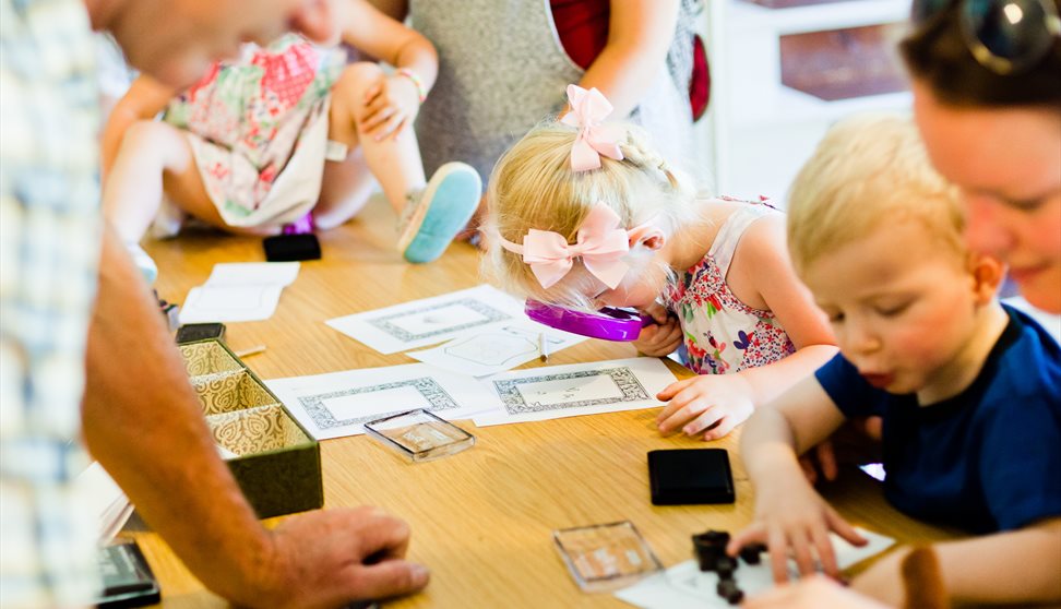 Children and adults gather around a table with papers, stamps, and a magnifying glass during an activity session. Children and adults gather around a table with papers, stamps, and a magnifying glass during an activity session.