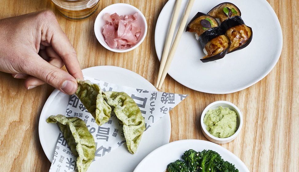 Small white plates of different foods displayed on a table with chopsticks and a hand holding a green gyoza.