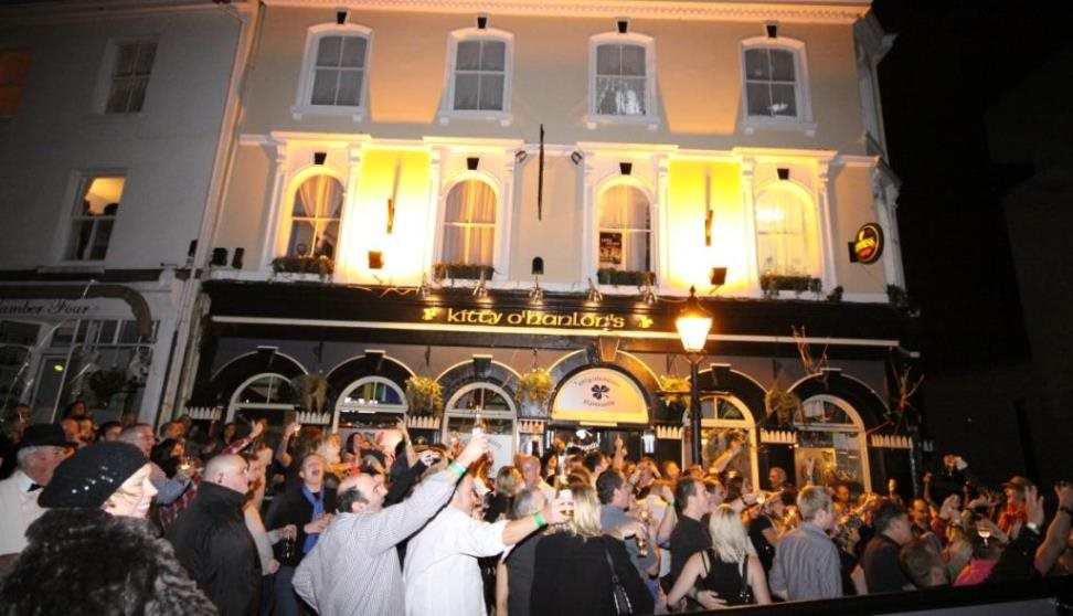 A crowd of people outside the brightly lit front of Kitty O'Hanlons at night-time.