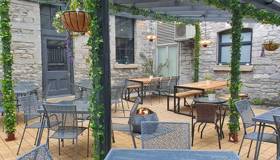 Outside courtyard area with tables and chairs, a fire pit and a plant covered pergola.