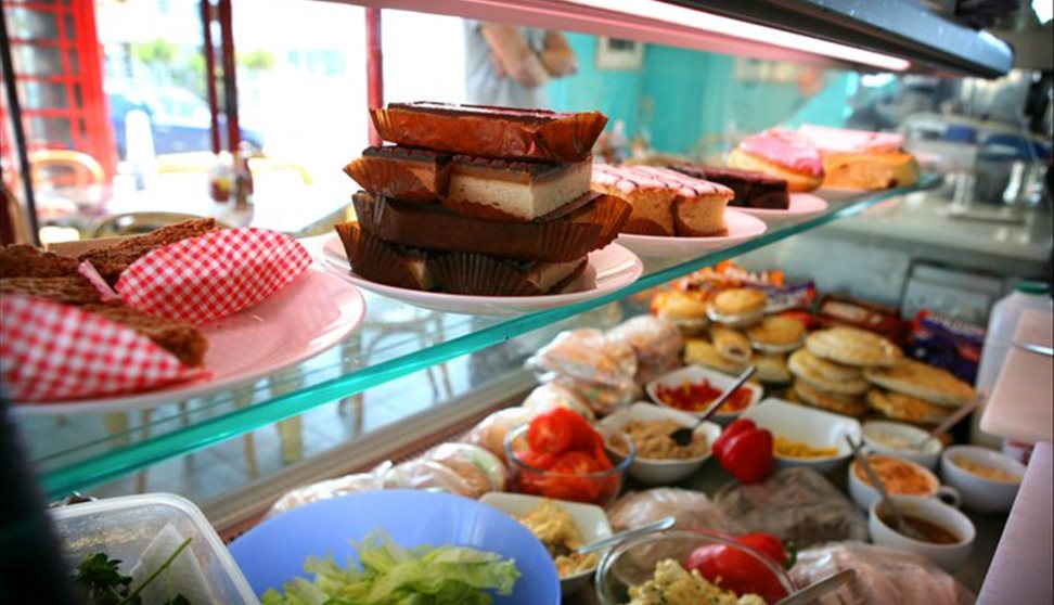 Large refrigerated display cabinet inside shop window displaying cakes, slices, pies and sandwich ingredients.