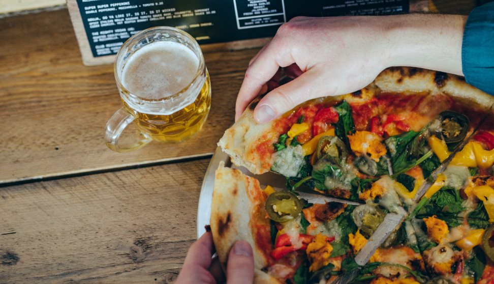 A large pizza divided into portions being pulled apart by a pair of hands. On the table is also a glass of beer and a menu.