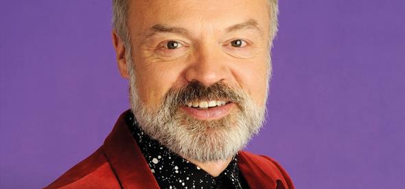Graham Norton in a red jacket and black button down against a purple backdrop