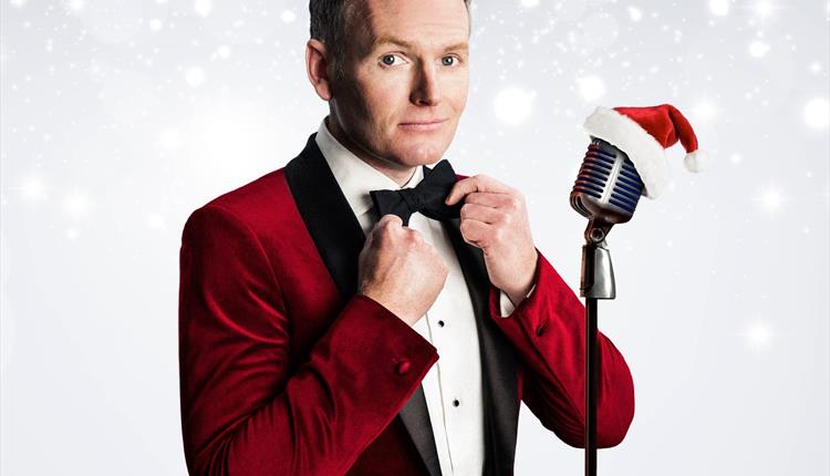 An image of singer Joe Stilgoe straightening a bow tie, whilst stood next to a microphone with a Santa hat placed on top.