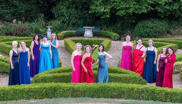 Choir of female singers, dressed in colourful evening gowns, standing in an ornamental garden with fresh greenery