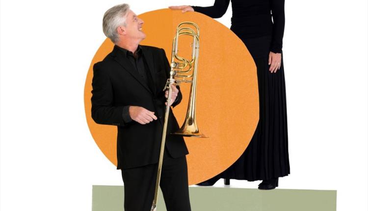 Image of a woman and a man both dressed in black against a white and orange background.  the man is holding a trombone.