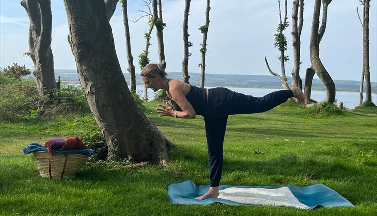 Yoga teacher in pose on a mat outside with trees and sea in the background.