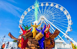 5 men playing instruments whilst dressed in colourful clothing in front of Bournemouths big wheel