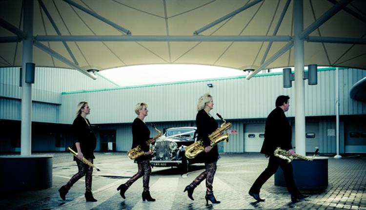 4 musicians carying various sizes of saxophone walking across a hangar building with a Rolls Royce in the background.