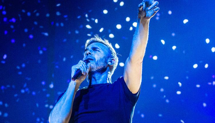Gary barlow points to the sky while singing infront of a blue background