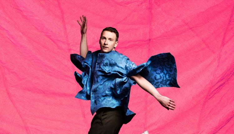 A man jumping mid air, with a flamboyant blue top on, against a pink background,