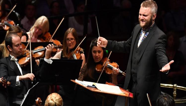 A conductor stood smiling at his orchestra with open arms as they play.