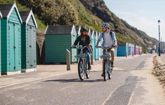 Couple cycling along Bournemouth seafront with beach huts behind them
