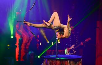 Image of a contortionist on stage shooting a bow and arrow with their feet amidst a colourful lit background