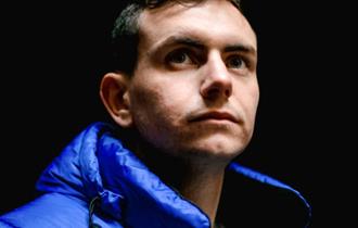 Image of the performer Connor Burns from the shoulders up in a blue puffer jacket against a black background