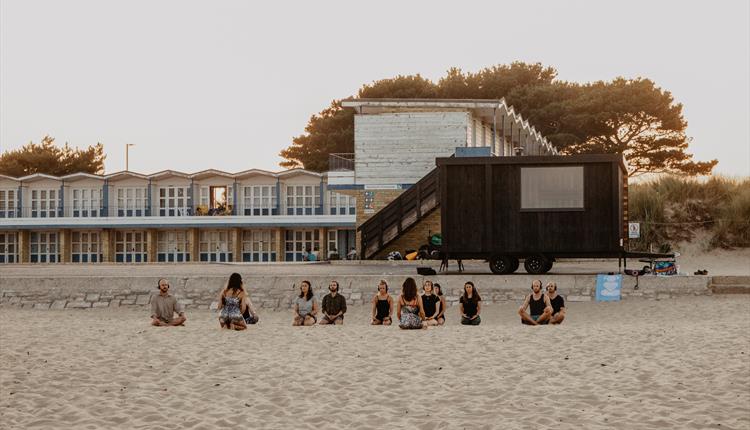 The Saltwater Sauna based at Sandbanks beach. A group of people sitting crossed legged on the sand in front of the sauna lodge.