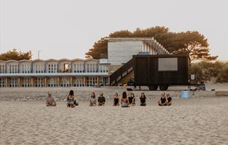 The Saltwater Sauna based at Sandbanks beach. A group of people sitting crossed legged on the sand in front of the sauna lodge.