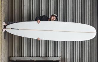 An image on its side of a man holding a long white surfboard infront of a corrugated iron shop front.