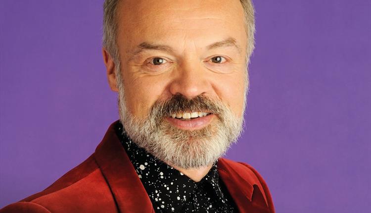Graham Norton in a red jacket and black button down against a purple backdrop