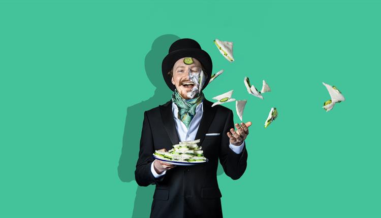 A man in a top hat, throws a plate of cucumber sandwiches in the air, some of which have landed on his face. He is smiling, the background is green.