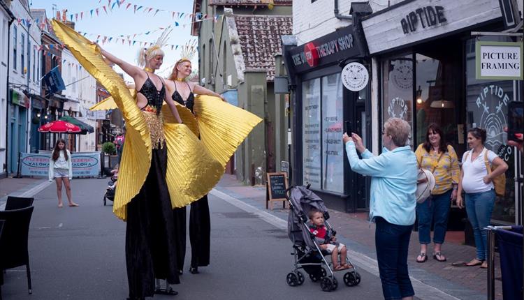 Two giant stiltwalkers dressed in black costumes with yellow capes posing to have their photo taken in Poole High Street