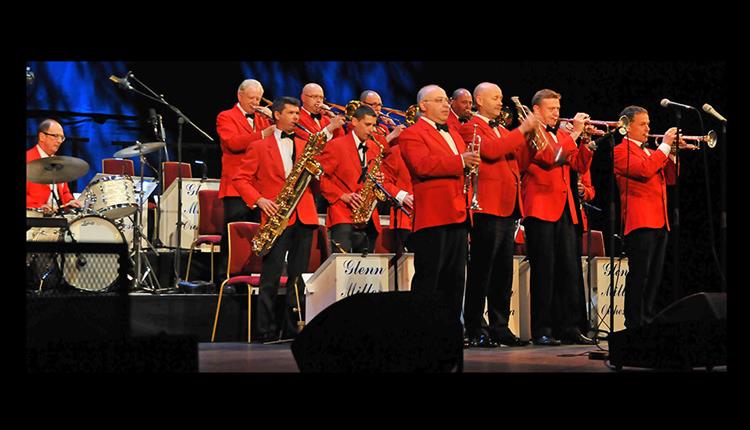 Glenn Miller Orchestra all standing and playing on stage wearing orange - red jackets
