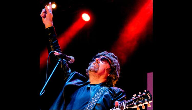 ELO, singer at mic, holding guitar and pointing up to ceiling whilst on stage performing. wearing sunglasses