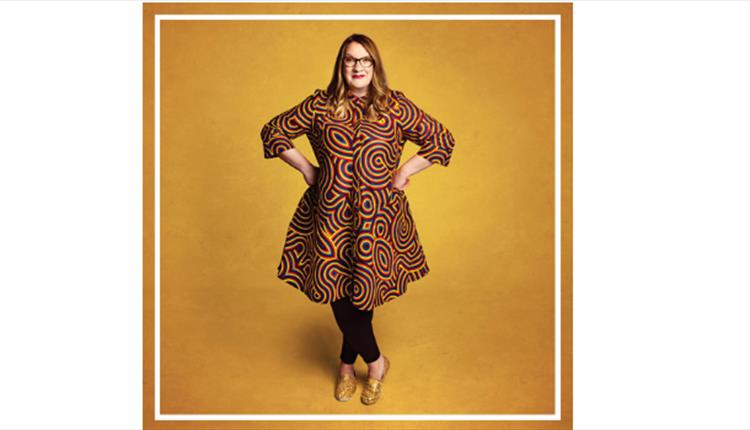 Sarah Millican standing with hands on hips smiling at camera wearing gold & brown dress