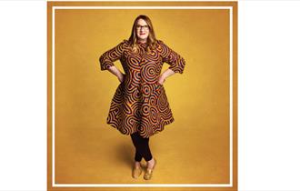 Sarah Millican standing with hands on hips smiling at camera wearing gold & brown dress