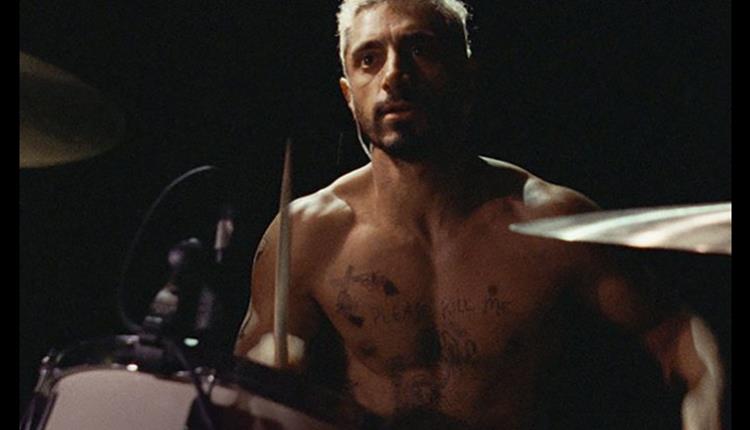 Topless man sits at drums with drumsticks in his hands.