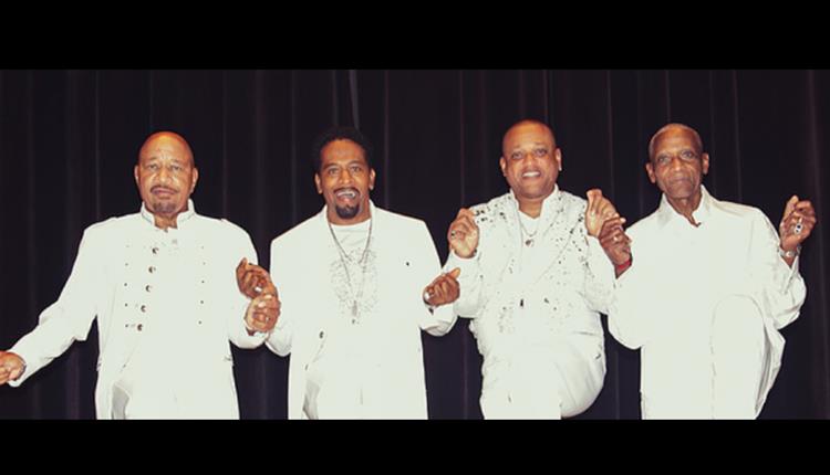 The Stylistics 4 male group in white jackets facing the camera and singing on stage with a dark background