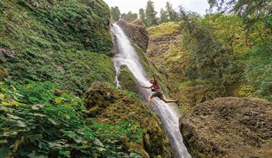Green landscape with a waterfall in the middle and a man jumping from a rock to a ledge.