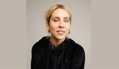 Poster image for Stevie Martin: Clout, showing the comedian in a black hoodie and stood against a brown background