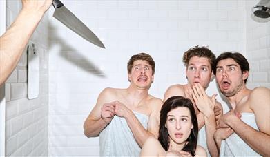 Three men, one woman in towels in a shower someone is holding a knife up out of shot and they look shocked.