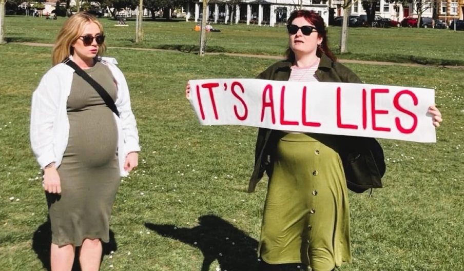 Two women on Southsea Common. One is pregnant, the other holds a white banner on which red text reads: It's all lies