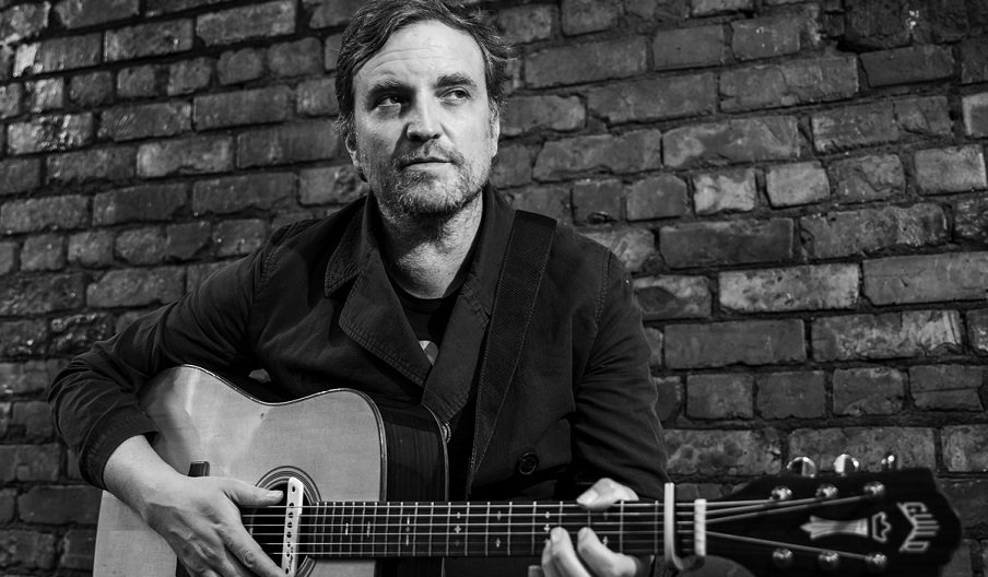 Black and white photograph of James Walsh with an acoustic guitar