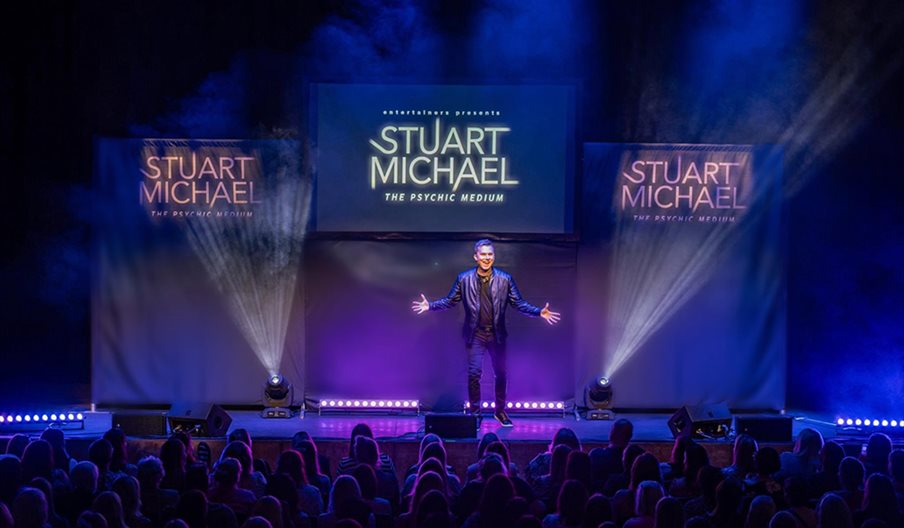 Stuart Michael stands on stage in front of an audience, there are three screens behind him with his name on.