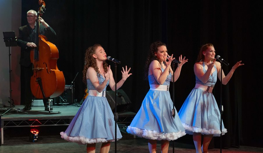 Three women dressed in blue 1950s style dresses singing with a cello player in the background.
