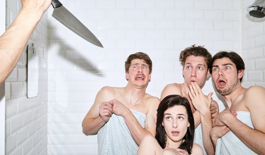 Three men, one woman in towels in a shower someone is holding a knife up out of shot and they look shocked.