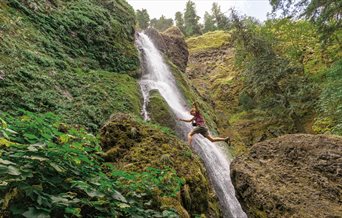 Green landscape with a waterfall in the middle and a man jumping from a rock to a ledge.