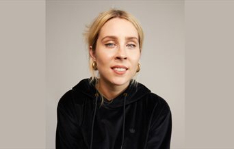 Poster image for Stevie Martin: Clout, showing the comedian in a black hoodie and stood against a brown background