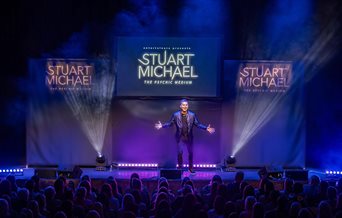 Stuart Michael stands on stage in front of an audience, there are three screens behind him with his name on.