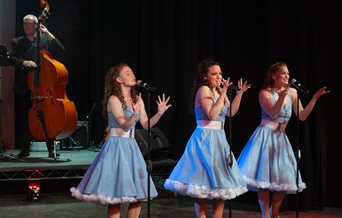 Three women dressed in blue 1950s style dresses singing with a cello player in the background.